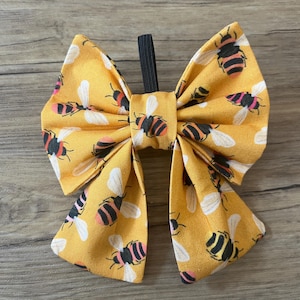 Peut inclure: Un nœud papillon en tissu jaune avec un motif répété d'abeilles noires, roses et blanches. Le nœud est attaché avec un élastique noir. Le nœud mesure environ 13 cm de large.