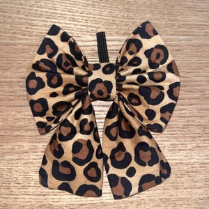 May include: A large bow with a leopard print pattern. The bow features a tan background with black and brown spots. The bow is attached to a black elastic band. The bow is made of a silky material and is likely used as a hair accessory.