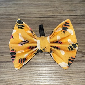 May include: A yellow bow tie featuring a pattern of black and yellow bees. The bow tie has a black elastic band. The fabric has a light yellow background with the bees in various positions.