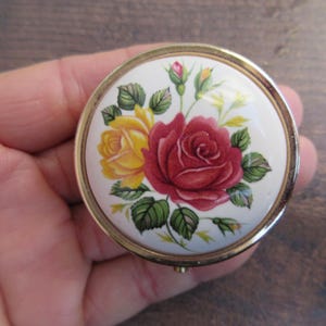 Può includere: Una piccola scatola rotonda con un bordo dorato e uno sfondo bianco. Il coperchio presenta un motivo floreale con rose rosse, gialle e verdi.