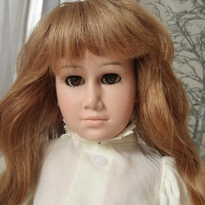 Puede incluir: Una muñeca de porcelana con el pelo largo y castaño peinado con raya lateral y flequillo. La muñeca lleva una blusa blanca con cuello de encaje y cierre de botón.