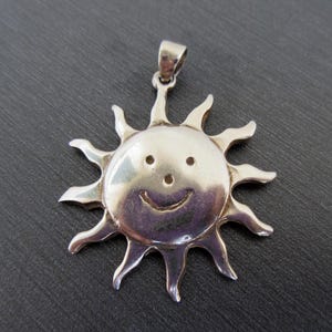 Könnte beinhalten: Silberfarbener Sonnenanhänger mit einem lächelnden Gesicht und strahlenden Strahlen. Der Anhänger hat oben eine Schlaufe für eine Kette. Das Gesicht der Sonne hat zwei kleine Punkte für Augen und eine gebogene Linie für ein Lächeln. Ein fröhliches Schmuckstück.