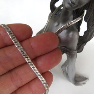 Op de afbeelding: Een zilverkleurige visgraatketting wordt in een hand gehouden, met een standbeeld van een vrouw op de achtergrond. De ketting heeft een plat, geweven ontwerp. Het standbeeld is grijs en heeft lang haar.