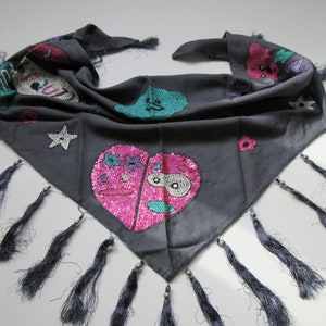 May include: A gray triangular scarf with black fringe and colorful embroidered designs. The designs include a heart with a face, a star, and a flower.