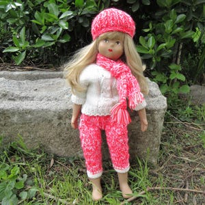 May include: A doll wearing a pink and white knitted hat, a white knitted sweater, and pink and white knitted pants. The doll has blonde hair and blue eyes.