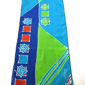 Può includere: Una lunga sciarpa rettangolare con un vivace design geometrico. La sciarpa presenta sezioni blu, verdi e arancioni, con motivi a spirale e quadrati bianchi. Il testo "Kaare Danish Design" è visibile in basso a destra.