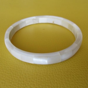 Può includere: Un braccialetto a cerchio bianco segmentato. Il braccialetto è composto da pezzi rettangolari di materiale bianco, creando un design circolare liscio. Il braccialetto è posto su uno sfondo giallo.