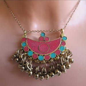 May include: A gold-toned necklace with a pendant featuring a pink and turquoise design. The pendant has a crescent shape with a central circle and is adorned with dangling gold beads. The necklace is displayed on a person.