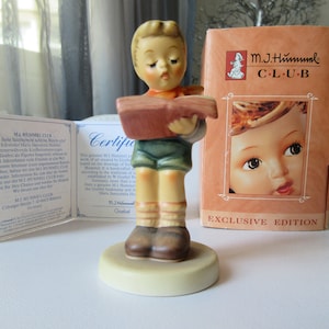May include: A porcelain figurine of a young boy holding a book, with a certificate and box. The figurine is part of the M.J. Hummel Club Exclusive Edition.
