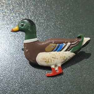 May include: A small, painted metal duck figurine. The duck is brown, green, white, and red. It has a long, thin beak and a small, round head.