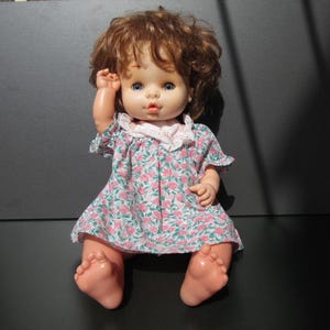 May include: A vintage plastic baby doll with brown curly hair, blue eyes, and a pink and green floral dress.