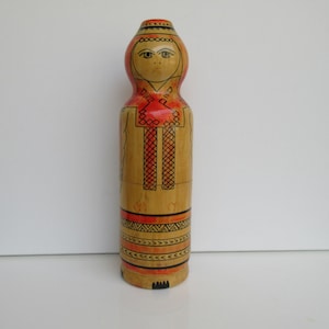 May include: A wooden doll with a painted face and traditional clothing. The doll is painted in shades of orange, yellow, and black.