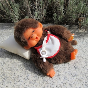 Puede incluir: Un muñeco de mono de peluche marrón con manos y pies naranjas, que lleva un babero blanco con ribete rojo y un chupete. El muñeco está tumbado sobre una pequeña almohada color crema. El mono tiene una expresión de sueño.