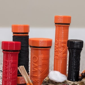 Peut inclure: Quatre contenants cylindriques orange et noirs avec le texte "TWO 5" et "ADDITIVE" imprimé dessus. Les contenants sont disposés sur une surface brune avec une boule de coton blanche, trois allumettes orange et deux piles rouges avec le texte "SURFIRE.COM" et "MADE IN USA" imprimé dessus.