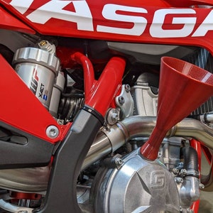 May include: Close-up of a red motorcycle engine with a silver exhaust pipe and a red funnel. The motorcycle has a white GASGAS logo on the side.