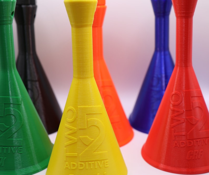 May include: Five colorful plastic bottles with a funnel shape and a narrow neck. The bottles are labeled with the text "TWO 52 ADDITIVE". The bottles are arranged in a row from left to right, with the green bottle in the front, followed by the black, yellow, red, and blue bottles.