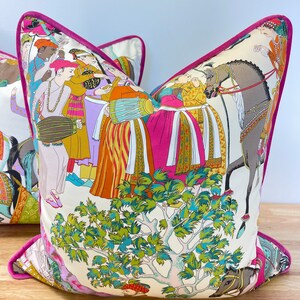 May include: A decorative pillow with a pink border and a white background featuring a colorful illustration of people, horses, and a tree. The illustration is in a traditional Indian style.
