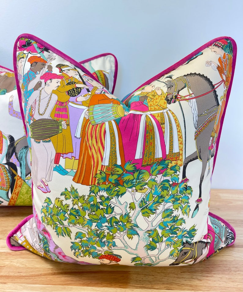 May include: A decorative pillow with a pink border and a white background featuring a colorful illustration of people, horses, and a tree. The illustration is in a traditional Indian style.