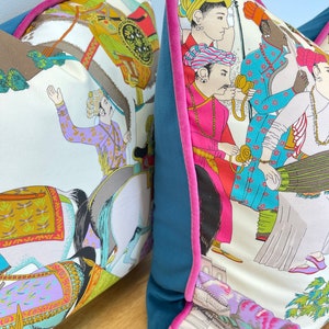 May include: Two decorative throw pillows with a white background and colorful illustrations of people riding horses and other figures. The pillows have teal blue velvet backing with a pink trim.