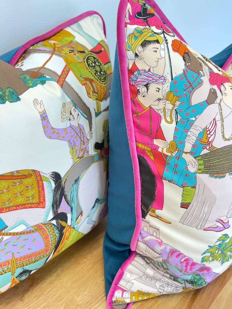 May include: Two decorative throw pillows with a white background and colorful illustrations of people riding horses and other figures. The pillows have teal blue velvet backing with a pink trim.