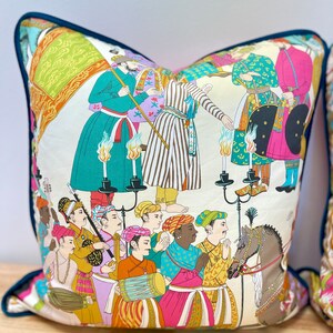 May include: A colorful throw pillow with a white background featuring a procession of people in traditional clothing. The scene includes musicians, a horse, and a group of people holding candles. The pillow has a teal blue border.