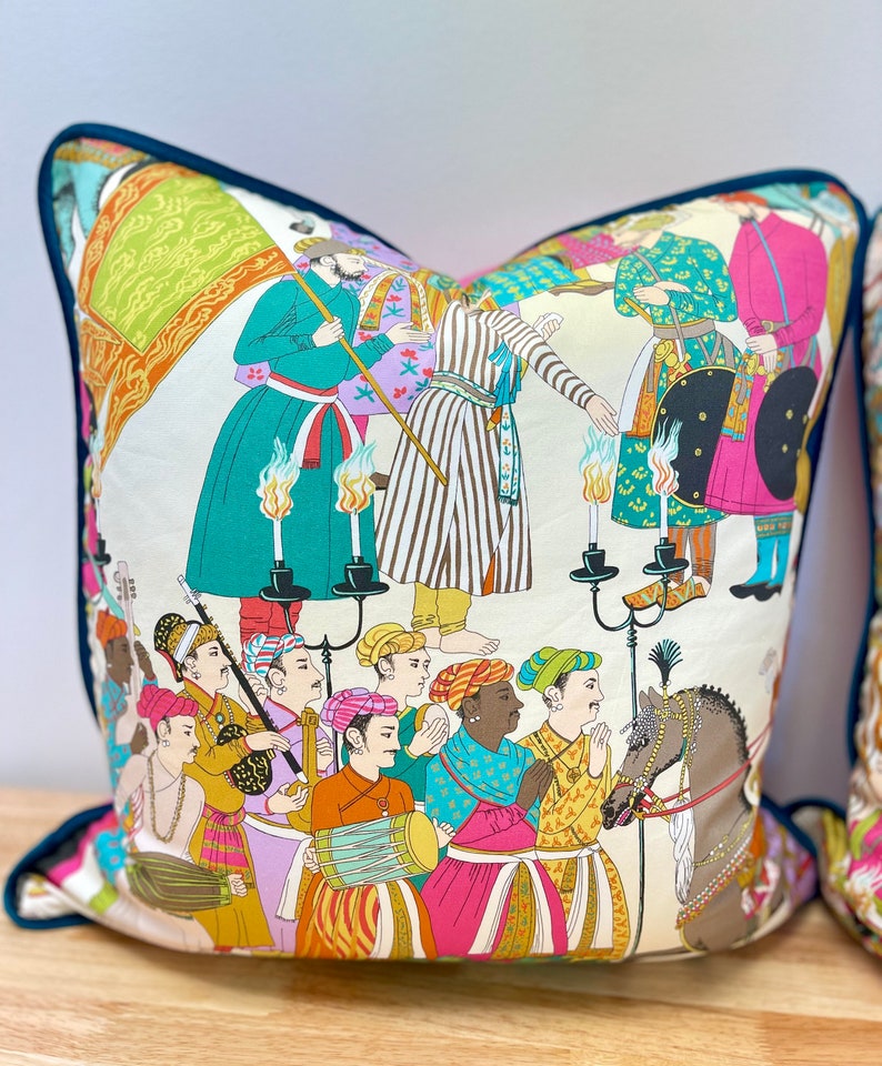 May include: A colorful throw pillow with a white background featuring a procession of people in traditional clothing. The scene includes musicians, a horse, and a group of people holding candles. The pillow has a teal blue border.