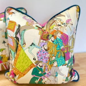 May include: A white pillow with a teal border features a colorful illustration of a procession of people on horseback. The people are wearing traditional clothing and turbans, and they are carrying flags and other objects. The background is a cream color.