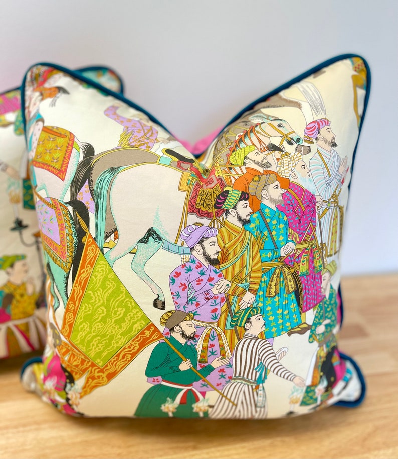 May include: A white pillow with a teal border features a colorful illustration of a procession of people on horseback. The people are wearing traditional clothing and turbans, and they are carrying flags and other objects. The background is a cream color.