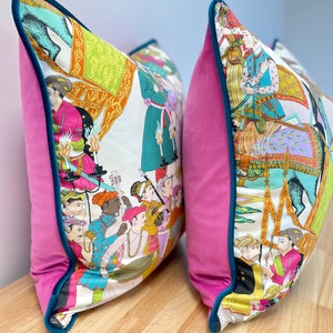 May include: Two decorative throw pillows with a pink velvet backing and a teal trim. The front of the pillows feature a white background with a colorful illustration of people riding elephants and playing music.
