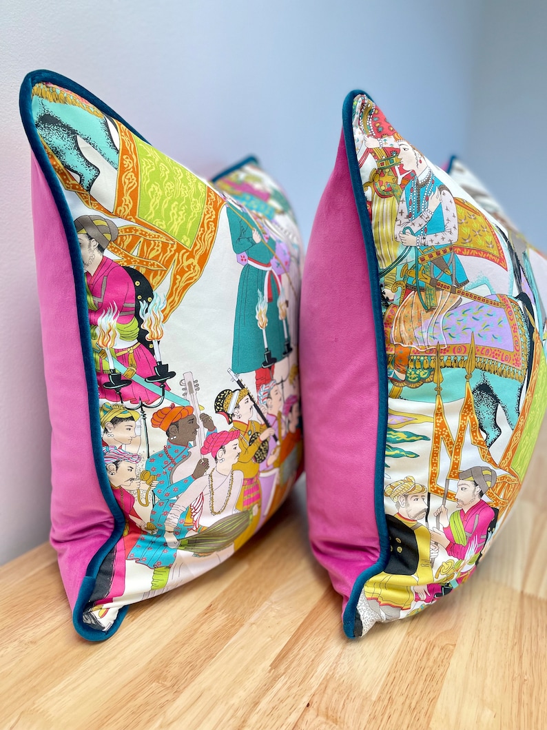 May include: Two decorative throw pillows with a pink velvet backing and a teal trim. The front of the pillows feature a white background with a colorful illustration of people riding elephants and playing music.