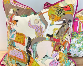 May include: A decorative pillow with a white background and a colorful design featuring people riding horses. The design is inspired by traditional Indian art and features intricate details and vibrant colors.