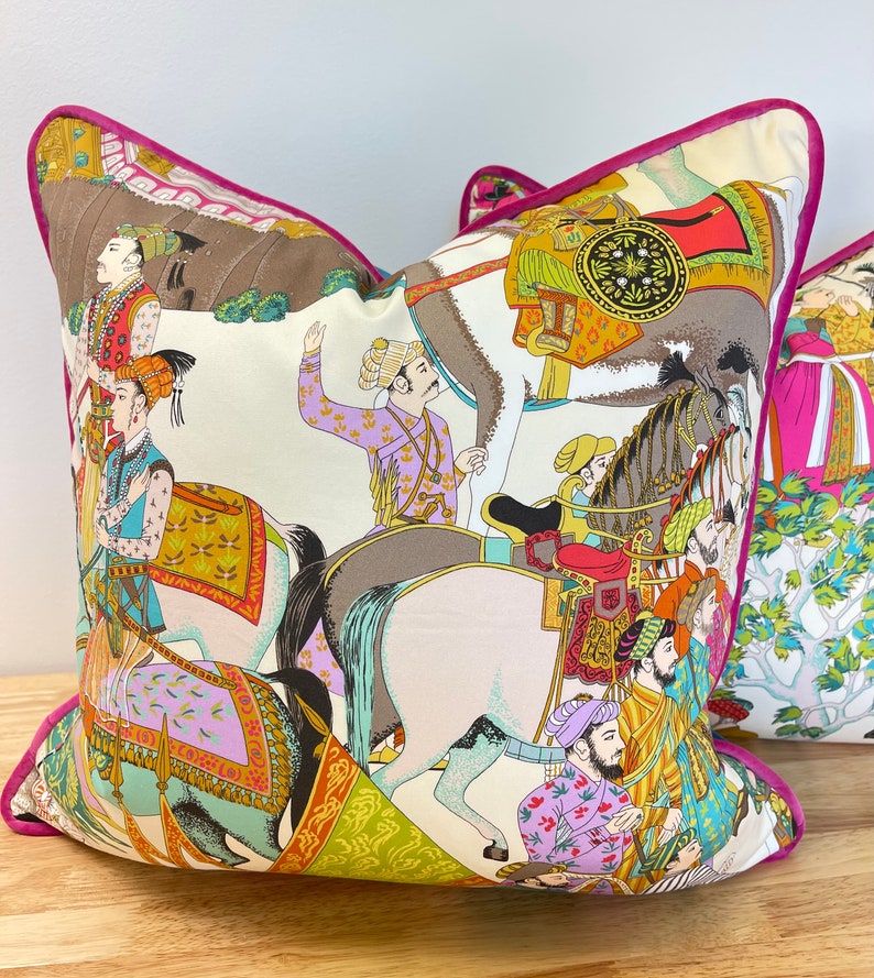 May include: A decorative pillow with a white background and a colorful design featuring people riding horses. The design is inspired by traditional Indian art and features intricate details and vibrant colors.