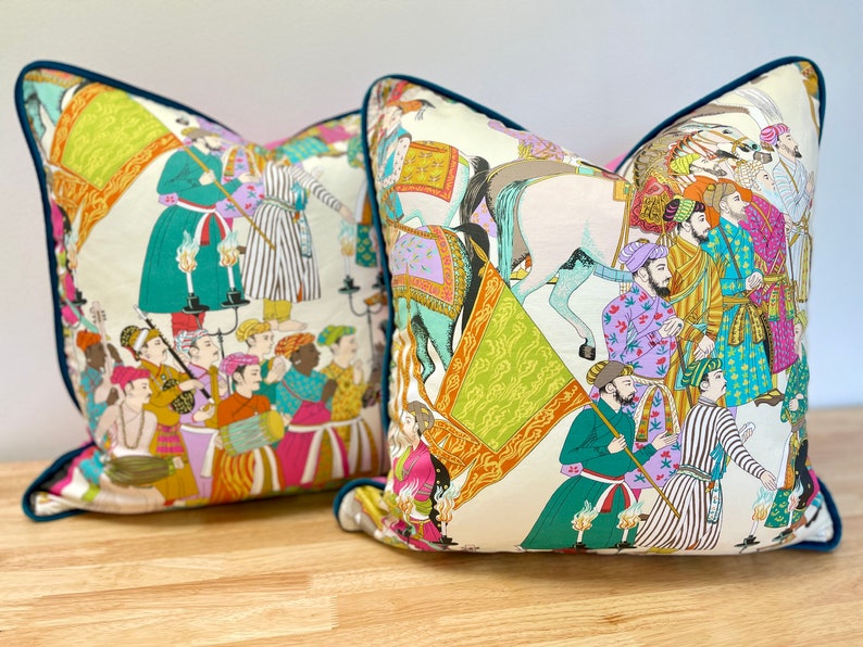 May include: Two decorative throw pillows with a white background and colorful illustrations of people and animals. The pillows have a teal blue trim.