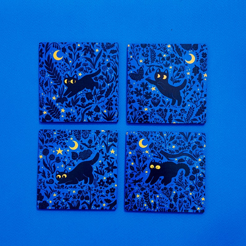 May include: Four blue square coasters with a black cat and moon design. Each coaster features a different cat pose and a crescent moon with stars.