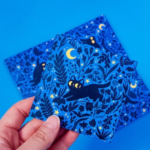 May include: Two blue cards with a black cat and floral pattern. The cards feature a black cat with yellow eyes walking through a field of flowers and stars. The text "Cloudy" is printed on one of the cards.