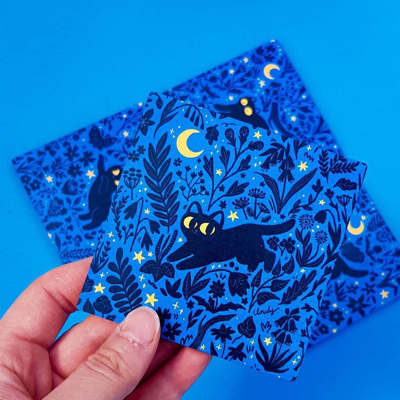 May include: Two blue cards with a black cat and floral pattern. The cards feature a black cat with yellow eyes walking through a field of flowers and stars. The text "Cloudy" is printed on one of the cards.