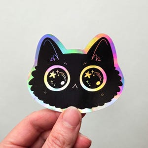 Op de afbeelding: Een zwarte kattensticker met grote, iriserende ogen met ster- en puntdetails. De kattenkop is omlijnd met een regenboog holografisch effect. De sticker wordt door een hand vastgehouden.