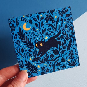 May include: A blue and black illustration of a cat with a crescent moon and stars on a dark blue background. The cat is surrounded by flowers and leaves.