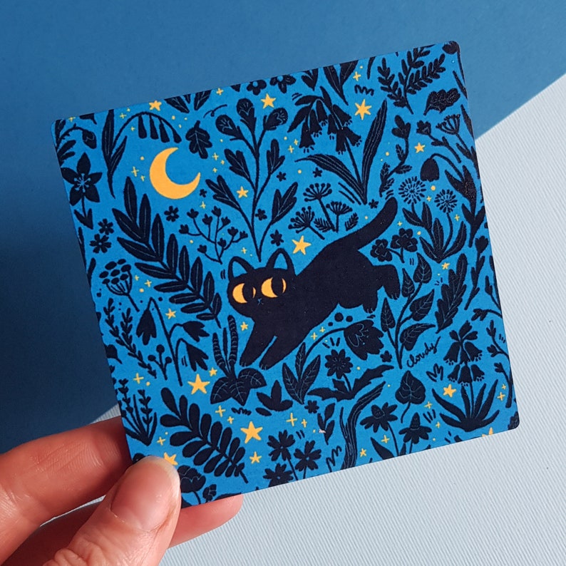 May include: A blue and black illustration of a cat with a crescent moon and stars on a dark blue background. The cat is surrounded by flowers and leaves.