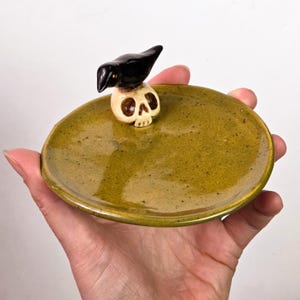 Może przedstawiać: Okrągła ceramiczna podstawka z błyszczącym, oliwkowozielonym nakrapianym szkliwem. Mały, trójwymiarowy czarny ptak siedzi na beżowej ludzkiej czaszce, która jest przymocowana do wewnętrznej krawędzi naczynia. Powierzchnia jest gładka i odblaskowa.