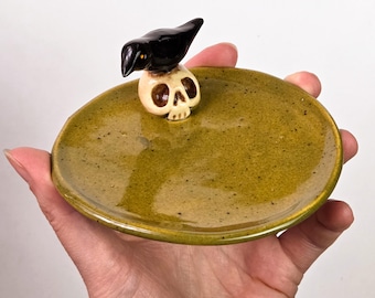 May include: A round ceramic trinket dish with a glossy olive green speckled glaze. A small, three-dimensional black bird is perched atop a beige human skull, which is attached to the inner edge of the dish. The surface is smooth and reflective.