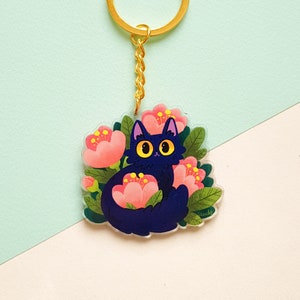 May include: A black cat with yellow eyes sits in a bed of pink flowers. The cat is surrounded by green leaves. The image is a keychain.