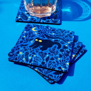 May include: Set of four blue coasters with a black cat design and yellow stars and flowers. The cat is walking through a forest with a crescent moon in the background.