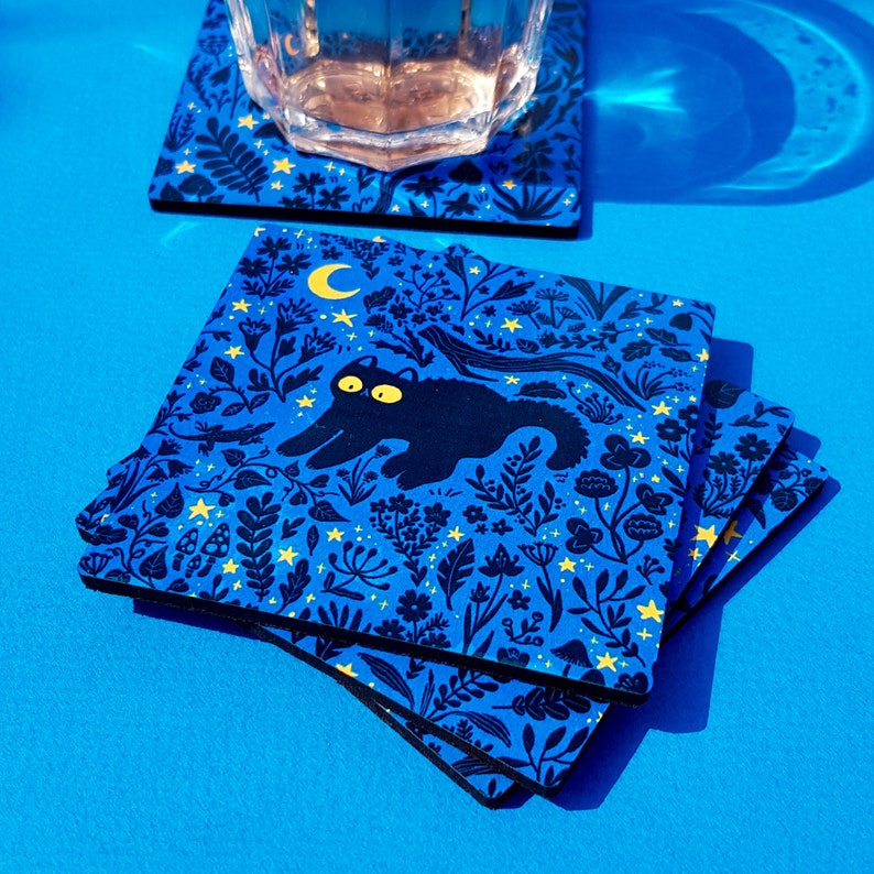 May include: Set of four blue coasters with a black cat design and yellow stars and flowers. The cat is walking through a forest with a crescent moon in the background.