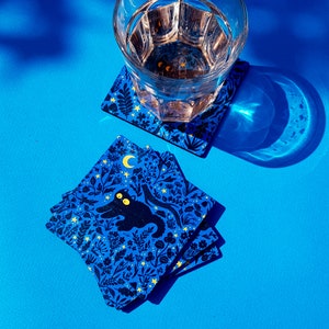 May include: Four blue coasters with a black cat and gold stars and moon design. The coasters are stacked on top of each other. A glass of water sits on top of the coasters.