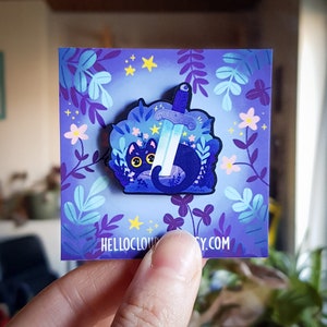 Könnte beinhalten: Dunkelblaue Emaille-Anstecknadel mit einer schwarzen Katze mit gelben Augen, einem Schwert und floralen Elementen. Die Nadel befindet sich auf einem lila Hintergrund mit floralem Design und dem Text "HELLOCLOUDSOCIETY.COM".