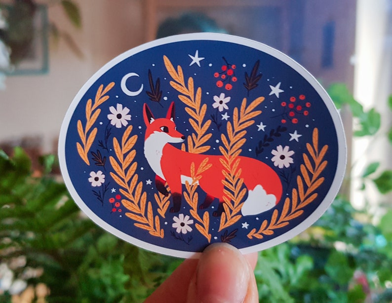 May include: A round sticker with a blue background featuring a red fox with a white tail tip surrounded by yellow and orange leaves, white flowers, and red berries. The sticker also has a crescent moon and white stars.