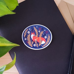May include: A round sticker with a blue background and a white border. The sticker features a fox sitting in a field of tall grass with white flowers and red berries. The fox is orange with a white chest and a black nose. There is a crescent moon and stars in the background.