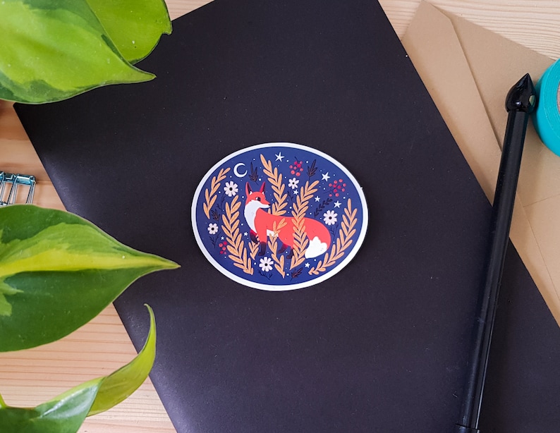 May include: A round sticker with a blue background and a white border. The sticker features a fox sitting in a field of tall grass with white flowers and red berries. The fox is orange with a white chest and a black nose. There is a crescent moon and stars in the background.