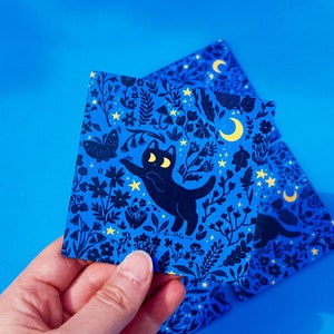 May include: A blue and black illustrated card featuring a black cat with yellow eyes and a crescent moon. The cat is surrounded by a variety of flowers and plants, all in a dark blue color. The card has a whimsical and magical feel.